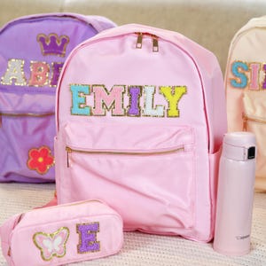 May include: Pink backpack with a gold zipper and a colourful felt name patch that says "EMILY". The backpack has a small front pocket and a matching pink pencil case with a butterfly and the letter "E" on it.