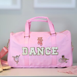 Op de afbeelding: Een roze danstas met het woord "DANCE" in witte pailletten letters. De tas heeft een schouderriem en handvatten en is versierd met ballet-thema applicaties. Balletschoenen en bloemen liggen naast de tas.