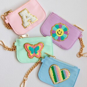Könnte beinhalten: Vier kleine Reißverschlusstaschen mit goldenen Ketten. Jede Tasche hat einen anderen Stoff und ein anderes gesticktes Design: ein weißer Buchstabe A, eine gelbe Blume mit einem Smiley-Gesicht, ein rosa und grüner Schmetterling und ein regenbogenfarbenes Herz.