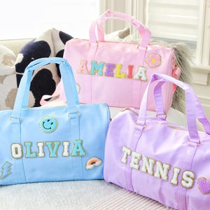 May include: Three personalised duffel bags in blue, pink and purple. The blue bag has the name "Olivia" on it, the pink bag has the name "Amelia" on it, and the purple bag has the word "Tennis" on it.