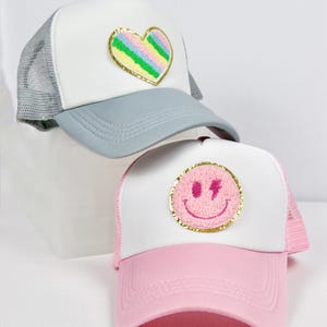 May include: Two trucker hats with embroidered patches. The first hat is grey with a white mesh back and a rainbow heart patch. The second hat is pink with a white mesh back and a pink smiley face patch with a lightning bolt.