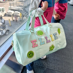Puede incluir: Una bolsa de lona azul claro con el nombre "ELLIE" en letras coloridas y brillantes. La bolsa presenta un parche de pelota de tenis, un rayo, una flor y una carita sonriente. La bolsa tiene dos asas y una correa para el hombro.