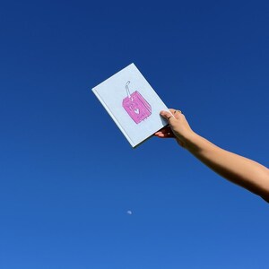 May include: A white notebook with a pink juice box illustration on the cover. The juice box has a straw and the words "Strawberry Milk" written on it. The notebook is held up against a bright blue sky.