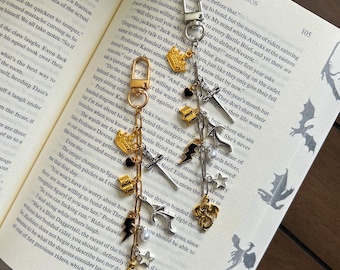 May include: Two decorative keychain charms with gold and silver tones, featuring dragon, star, and crown pendants. The charms hang against an open book with text and illustrations, creating a fantasy theme.