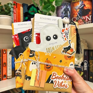 May include: A Halloween-themed gift set wrapped in orange paper with ghost and pumpkin patterns. The package is tied with twine and includes a ghost-shaped item, reading lists, and stickers that say "Bookish Vibes".