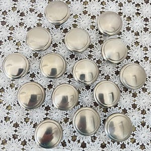 May include: A collection of silver-toned, round cabinet knobs. Each knob has a slightly domed top and a circular base. The knobs are arranged on a white lace surface, creating a vintage aesthetic. The knobs are likely for furniture restoration or DIY projects.