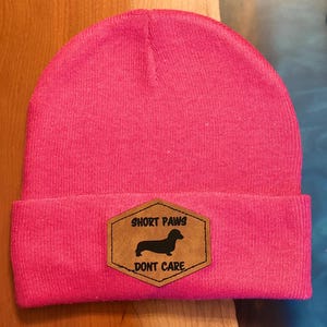 May include: A vibrant pink beanie hat with a turned-up hem. A brown patch displays a black dachshund illustration and the text "SHORT PAWS DONT CARE".