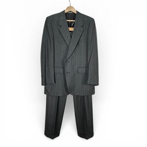 May include: A gray pinstripe suit, including a blazer and trousers, hangs on a wooden hanger. The blazer has two front pockets and a notched lapel. The trousers have a classic cut with a cuffed hem. The suit is made of a textured fabric.