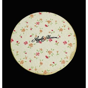 May include: A round, cream-colored container with a floral pattern of pink and peach roses and green leaves. The words "Apple Blossom" are written in cursive script across the center. The container has a gold-colored rim.