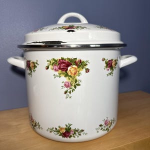 Könnte beinhalten: Weißer Emaille-Topf mit Deckel, verziert mit einem floralen Rosenmuster in Rot, Gelb und Rosa. Der Topf hat zwei seitliche Griffe und einen Deckelgriff. Der Rand ist silberfarben.