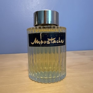 Peut inclure: Flacon de cologne en verre transparent avec un bouchon argenté. La bouteille présente des rainures verticales et contient un liquide jaune clair. Une étiquette bleu marine avec des lettres dorées indique "Moustache E Cologne."
