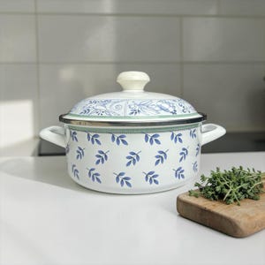 May include: White enamel cooking pot with a lid, decorated with blue leaf patterns and a floral design on the lid. The pot has two handles and a silver rim. A small wooden board with herbs is next to the pot.