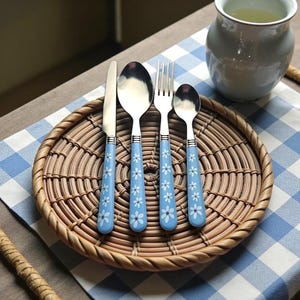Puede incluir: Un juego de cubiertos de acero inoxidable con mangos azul claro decorados con motivos florales blancos. El juego incluye un cuchillo, una cuchara, un tenedor y una cucharadita, dispuestos sobre un plato de mimbre tejido. Una jarra blanca y un paño a cuadros azul y blanco están en el fondo.