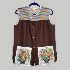 May include: A sleeveless brown apron with a striped top in shades of red, yellow, blue, and green. The apron has two pockets with floral basket designs and white trim. It is buttoned down the front and hangs on a blue hanger.