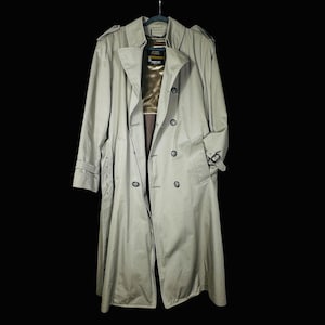 May include: A light tan trench coat with a double-breasted design, hanging on a black hanger. The coat features a notched collar, button closures, and a belt detail. The coat's length extends to the ankles.