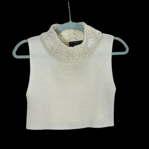 May include: Cream-colored sleeveless sweater with a ribbed knit body and a decorative, open-knit cowl neck. The sweater is hanging on a light blue hanger against a black background. The label reads "Specialty Knits".