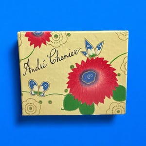 May include: A rectangular box with a cream-colored background featuring a large red flower, green leaves, and two blue and white butterflies. The text André Chenier is written in black cursive script on the left side of the box, surrounded by decorative line-drawn floral patterns.