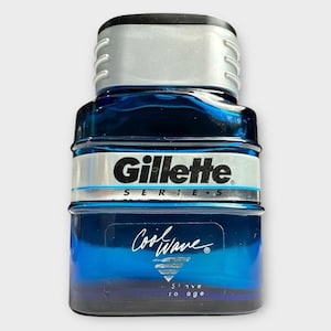 Puede incluir: Una botella azul de aftershave Gillette Series Cool Wave. La botella tiene una tapa plateada y una etiqueta plateada y negra con el logotipo de Gillette. Las palabras "Cool Wave" están escritas en cursiva en la parte delantera de la botella.