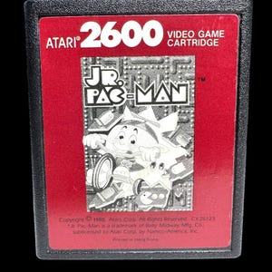 May include: Atari 2600 video game cartridge for Jr. Pac-Man. The cartridge has a red label with the Atari logo and the game title. The game's graphics feature a cartoon Pac-Man character. The cartridge is black.