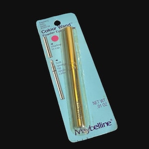 May include: A Maybelline Colour Wand powder eye shadow in a gold-toned cylindrical case, packaged on a light blue card. The packaging displays the text Raspberry Ruby (pearl) 425DS-04, Shadow in cap, Soft sponge tip applicator, and NET WT .01 OZ.
