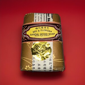 May include: A rectangular bar of Bee & Flower Sandal Wood Soap, wrapped in brown paper with a decorative label. The label features the brand name and product details. A gold foil seal is affixed to the front. Net weight is 2.65 oz.