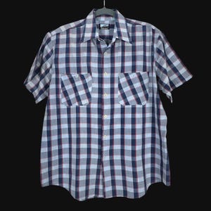 May include: A short-sleeved button-down shirt with a blue, white, and red plaid pattern. The shirt has two chest pockets and a classic collar. The garment is hanging on a hanger against a dark background.