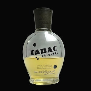 May include: A clear, rounded glass bottle of Tabac Original Eau de Cologne with a dark cap. The bottle contains a yellow liquid and features black text reading "TABAC ORIGINAL" and "EAU de COLOGNE".