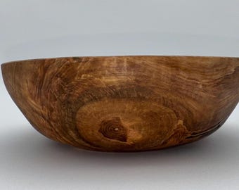 May include: A handcrafted wooden bowl with a natural, warm brown color. The bowl's interior and exterior showcase the wood grain, with a prominent knot visible at the base. The bowl has a smooth, rounded shape, perfect for serving or display.