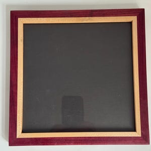 May include: A square picture frame featuring a deep purple outer border and a lighter tan inner border surrounding a solid black center panel. The frame has a clean, flat design with visible wood grain textures on both the purple and tan sections.