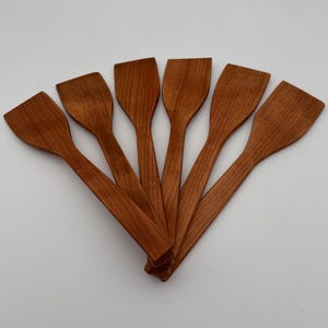May include: Six wooden spatulas fanned out against a white background. The spatulas are made of a reddish-brown wood, with a flat, rectangular head and a long, slender handle. The wood grain is visible.