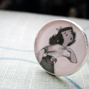 May include: A silver ring with a white cameo featuring a black and white image of a woman with short hair wearing a white shirt and a black belt.