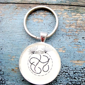 May include: A silver key chain with a round, white charm featuring a black and white design of a stylized face with a geometric pattern.