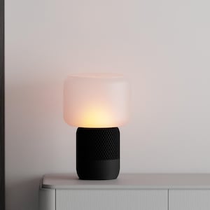 Könnte beinhalten: Eine moderne Tischlampe mit einem Schirm aus mattiertem, weißem Glas und einem schwarzen Sockel. Die Lampe strahlt ein warmes, einladendes Licht aus. Der Sockel hat ein strukturiertes, geometrisches Muster. Die Lampe steht auf einer hellgrauen Oberfläche.