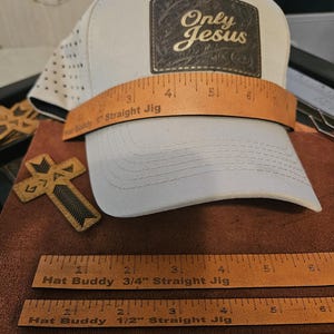 Może przedstawiać: Jasnoszara czapka z daszkiem z ciemnobrązową prostokątną naszywką z napisem Only Jesus. Na przodzie czapki umieszczono narzędzie pomiarowe Hat Buddy Straight Jig o szerokości 1 cala, a poniżej znajdują się szablony 3/4, 1/2 i 1/4 cala.