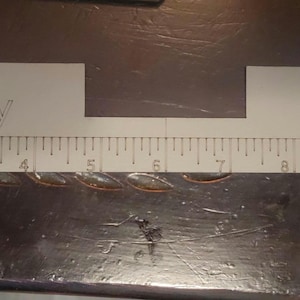 May include: A white rectangular measuring tool with a cutout section in the center. The text Hat Buddy is printed on the left side and 2.5" Square is printed on the right. A 12-inch ruler scale is printed along the bottom edge of the tool.