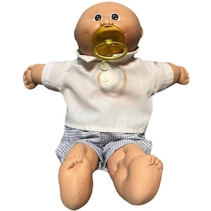 May include: A vintage Cabbage Patch Kid doll with a bald head, brown eyes, and a pacifier. The doll is wearing a white collared shirt, blue and white checkered shorts, and has tan-colored arms and legs.