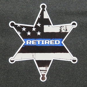 May include: A star-shaped badge with a white outline, featuring a distressed American flag design in black, white, and blue. The word "RETIRED" is displayed in white letters on a blue stripe across the center. The badge has a vintage, worn appearance.
