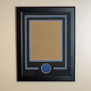 May include: A black picture frame with a double mat in white and blue, surrounding a tan-colored backing. The frame has a rectangular opening and a circular blue accent at the bottom.