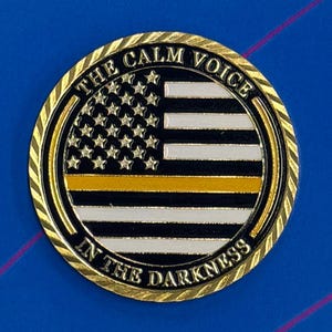 May include: A circular gold-toned metal coin featuring a stylized American flag with black, white, and a single horizontal yellow stripe. The text THE CALM VOICE IN THE DARKNESS is embossed in gold lettering along the outer rim of the coin.