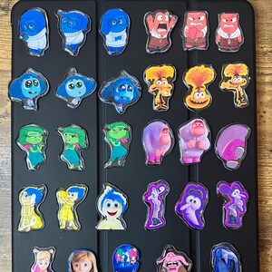 May include: A collection of colorful character charms from the animated movie "Inside Out". The charms feature various emotions, including Joy, Sadness, Anger, Fear, and Disgust, in a variety of poses. The charms are arranged on a black surface.