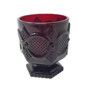 Pode incluir: Um cálice de vidro vermelho profundo com um exterior texturizado, apresentando padrões de diamante e motivos circulares em relevo. O recipiente tem uma abertura larga e assenta sobre uma base espessa de formato quadrado com bordas.