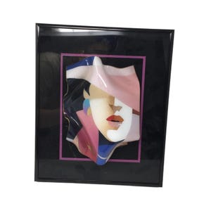 May include: Abstract art piece featuring a stylized face with a pink hat, red lips, and blue and white accents. The artwork is framed in a black frame with a pink border, creating a modern and colorful aesthetic.