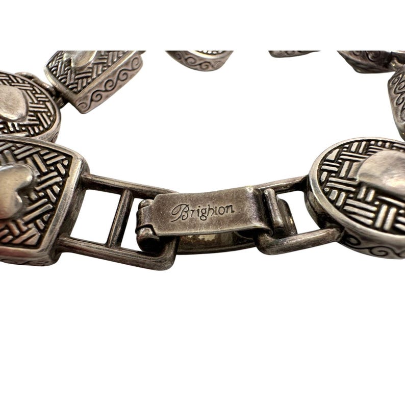 K&ouml;nnte beinhalten: Nahaufnahme eines silberfarbenen Armbands mit dekorativem Design. Das Armband hat einen Verschluss mit dem eingravierten Markennamen "Brighton". Die Armbandglieder haben ein strukturiertes Muster und herzf&ouml;rmige Akzente.