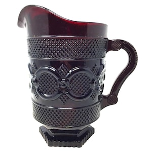 Puede incluir: Una jarra de vidrio rojo rubí con un diseño decorativo. La jarra presenta un diseño tallado en forma de diamante alrededor de la parte superior e inferior, con un intrincado patrón floral y ovalado en el centro. Tiene un asa y un pico para verter.