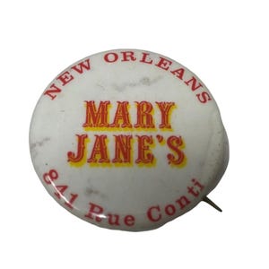 May include: A white button with red and yellow text. The text reads "New Orleans Mary Jane's 841 Rue Conti".