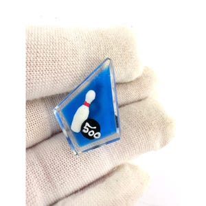 May include: A small, clear, triangular-shaped pin with a blue background. The pin features a white bowling pin with a red stripe and the number "500" in black. The pin is likely a collectible or a piece of jewelry.