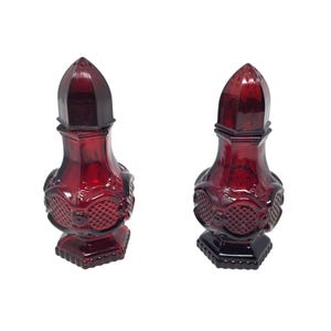 Puede incluir: Dos saleros y pimenteros de vidrio rojo rubí. Cada uno tiene una parte superior facetada y puntiaguda y un cuerpo decorativo texturizado. La base cuadrada tiene un patrón detallado. Los saleros y pimenteros están sobre un fondo blanco.