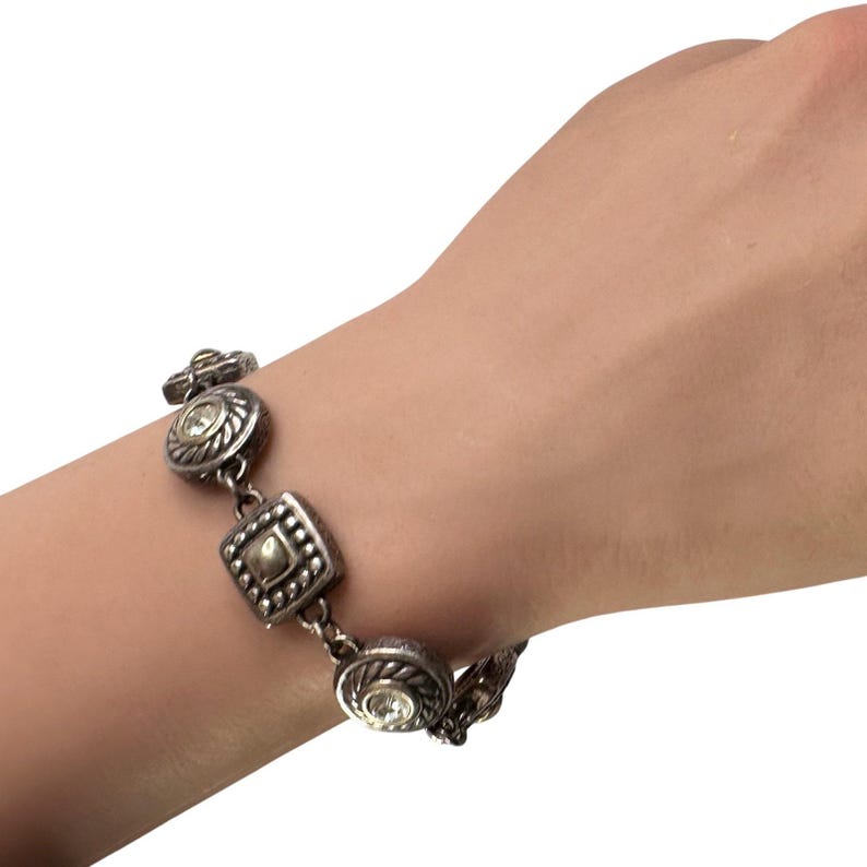 K&ouml;nnte beinhalten: Silberfarbenes Armband mit dekorativen Gliedern. Das Armband hat runde und quadratische Glieder mit aufwendigen Details und klaren, runden Edelsteinen. Das Armband wird am Handgelenk getragen.