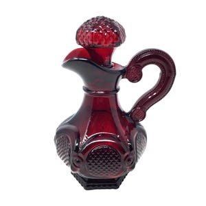 Puede incluir: Una jarra de vidrio rojo rubí con asa y tapón texturizado. La jarra tiene un pico y un asa decorativa. El cuerpo de la jarra presenta un patrón de diamantes y diseños circulares. Probablemente se utiliza para servir líquidos.