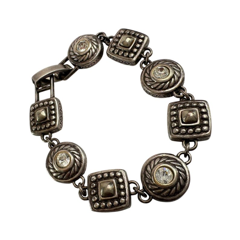 K&ouml;nnte beinhalten: Silberfarbenes Armband mit einer Mischung aus runden und quadratischen Gliedern. Jedes Glied hat einen erhabenen, strukturierten Rand und einen zentralen, klaren Kristall. Das Armband ist mit kleinen Gliedern und einem sicheren Verschluss verbunden.